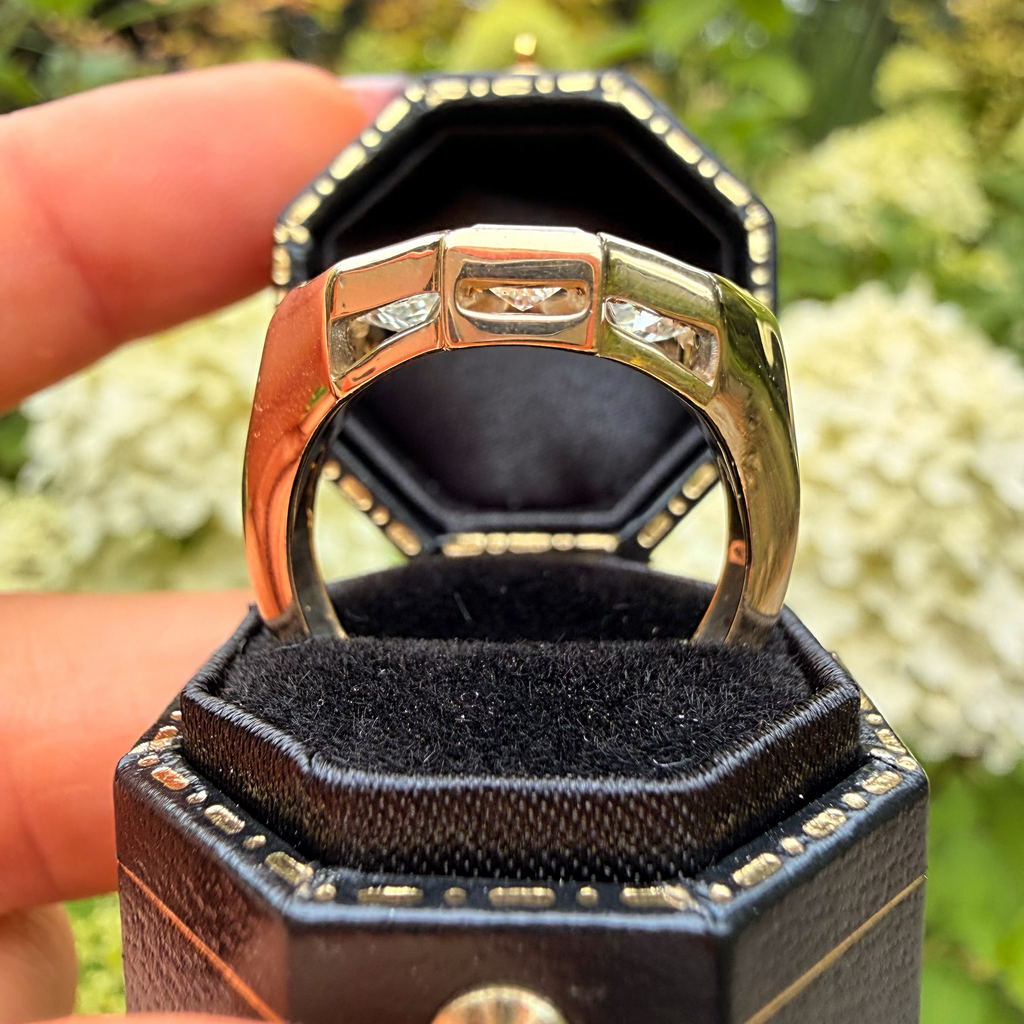 A close-up of a gold-toned ring displayed in an open black jewelry box held by a hand, with a blurred garden background of green foliage and white flowers.