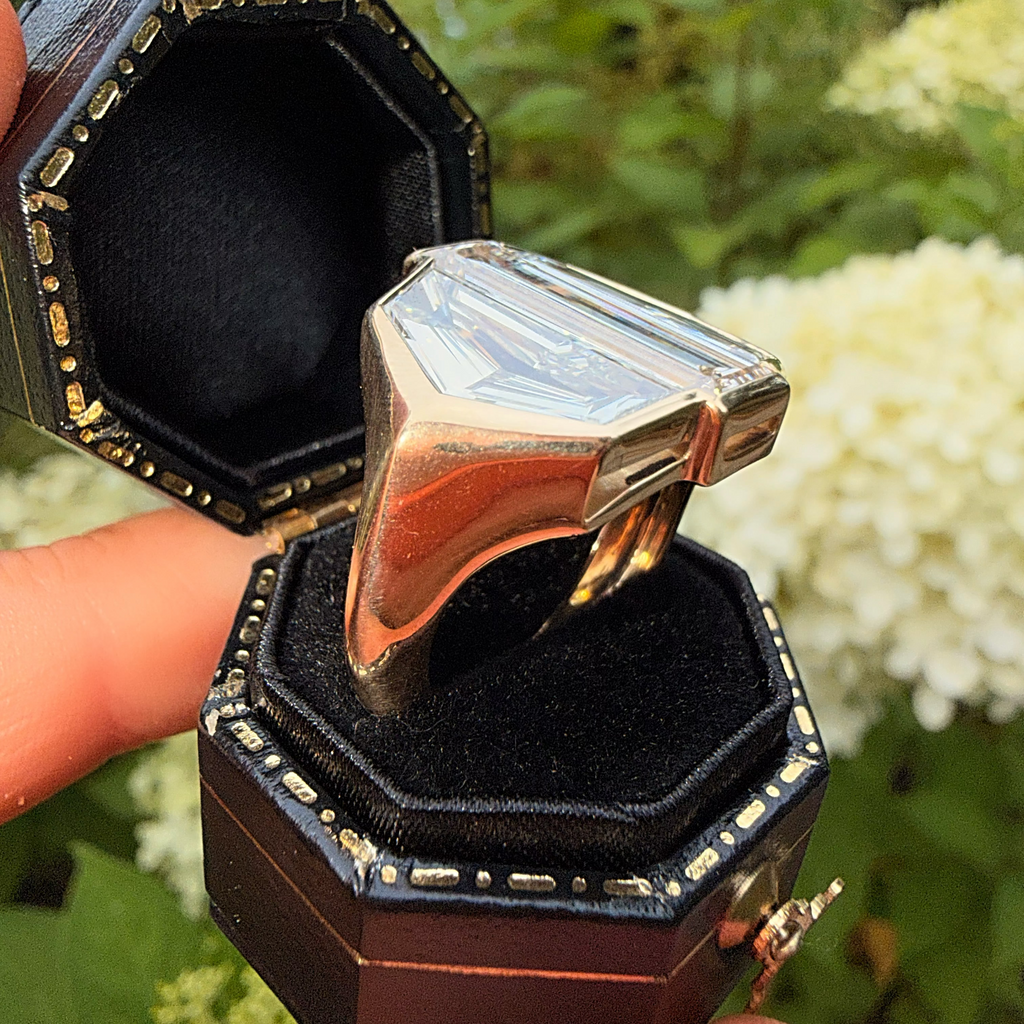 A large geometric silver ring displayed in an open dark brown octagonal ring box held by hands, with a blurred green garden background.