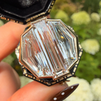 A close-up of a hand holding an open octagonal ring box with a large clear baguette-cut gemstone in a gold-toned setting, with blurred green foliage and white flowers in the background.