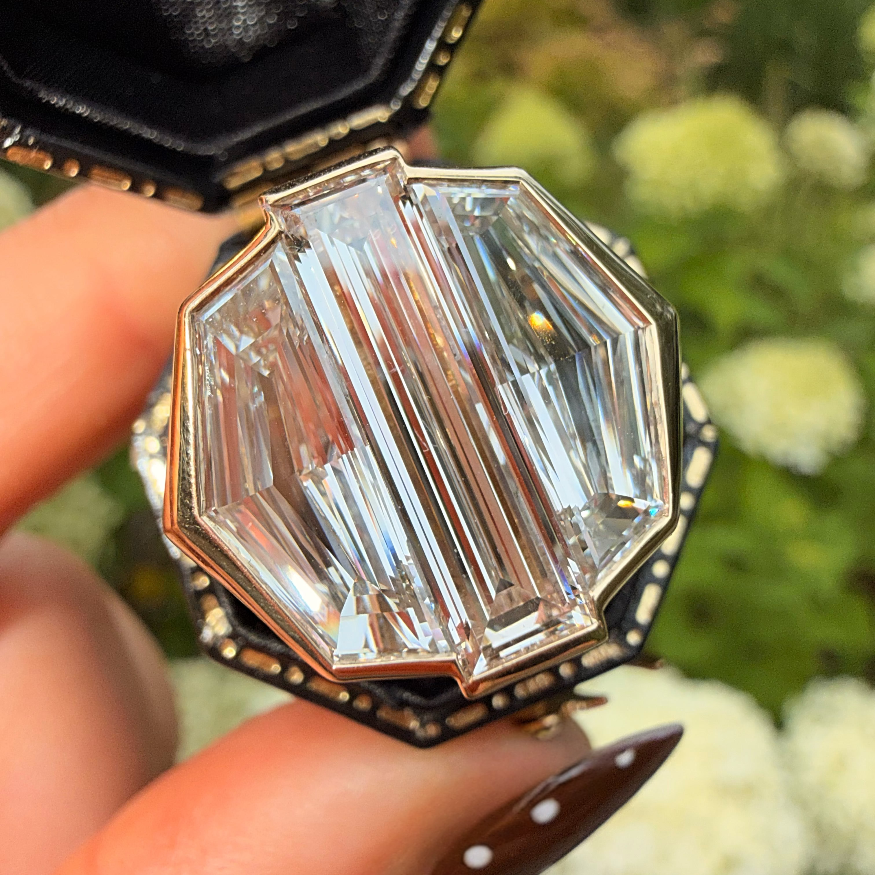 A close-up of a hand holding an open octagonal ring box with a large clear baguette-cut gemstone in a gold-toned setting, with blurred green foliage and white flowers in the background.