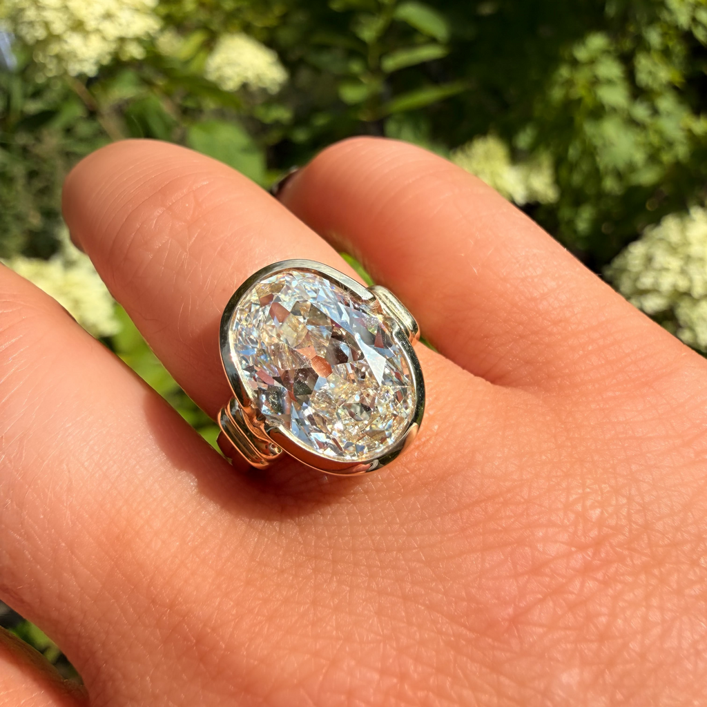 A gold ring with a large, round, iridescent clear gemstone worn on a person's finger, set against a blurred background of green foliage and white flowers.