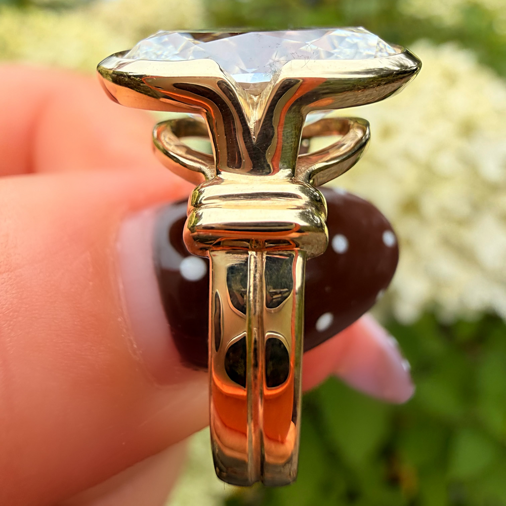 A gold ring with a large iridescent, color-shifting gemstone held by a hand with dark brown nail polish against a blurred outdoor background of green foliage and white flowers.