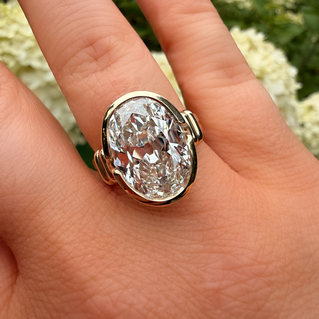 A close-up of a hand wearing a large oval diamond ring with a gold setting, with green foliage and white flowers blurred in the background.