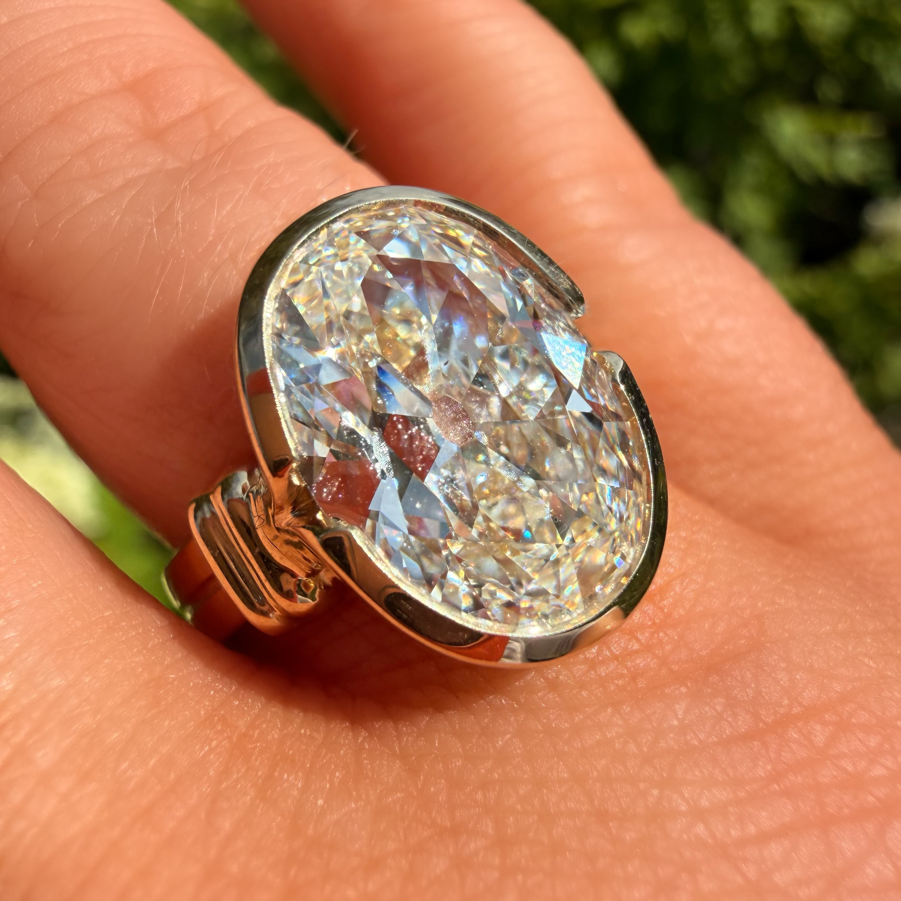 A close-up of a large oval-cut clear gemstone ring set in a gold-toned band, worn on a person's finger with a blurred green outdoor background.