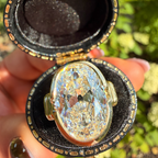 A large oval clear gemstone ring displayed in an open black and gold keepsake box held by a hand, with a blurred green outdoor background.