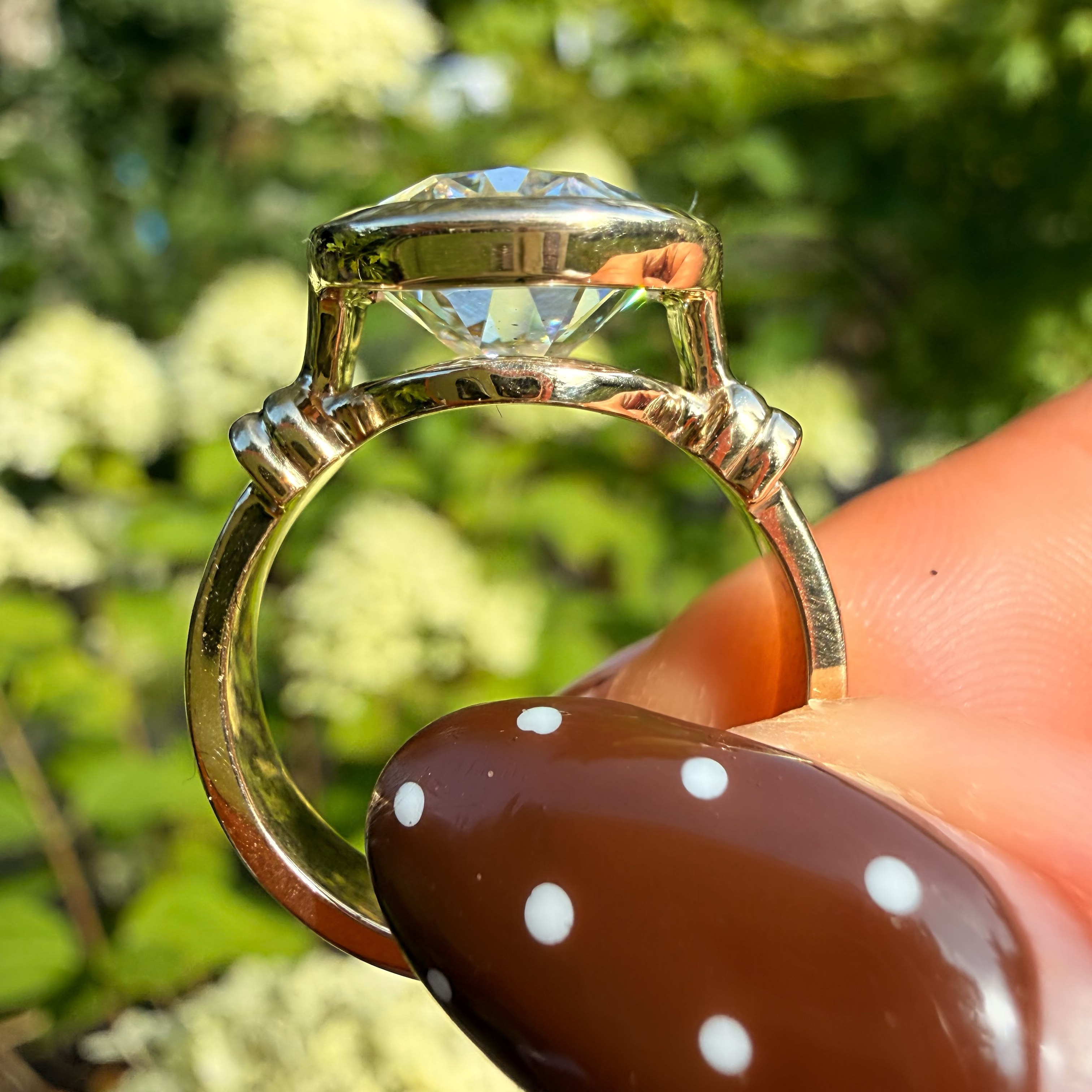 A gold ring with a large clear gemstone held by fingers with a brown and white polka dot nail design, set against a blurred green garden background.