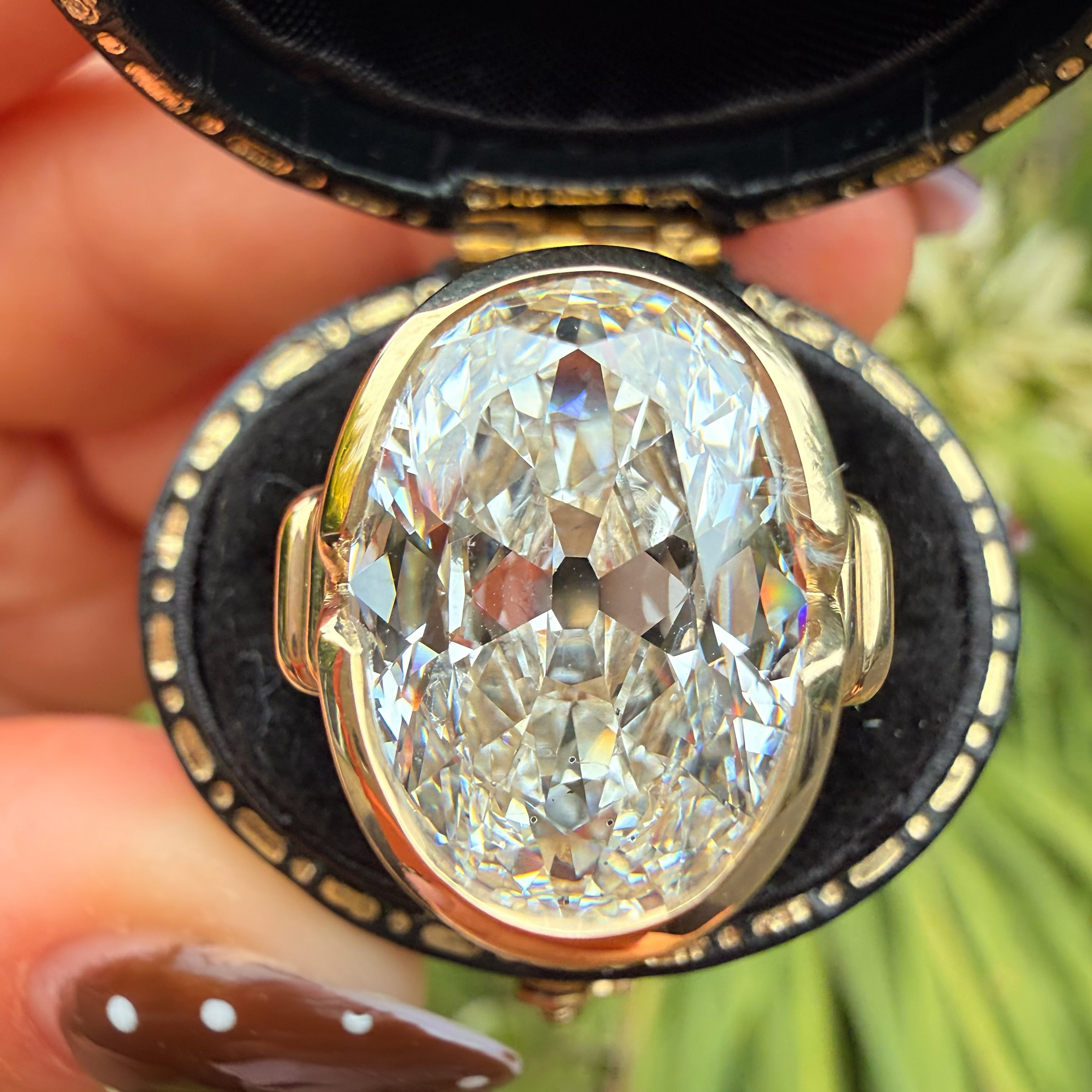 A close-up of a large oval clear gemstone set in a gold-toned ring, held inside an open black jewelry box by a hand with brown nail polish.