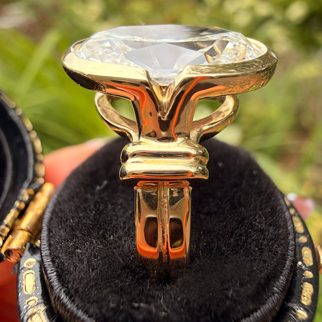 A gold ring with a large clear faceted gemstone displayed in an open black velvet ring box, held by a hand against a blurred green outdoor background.