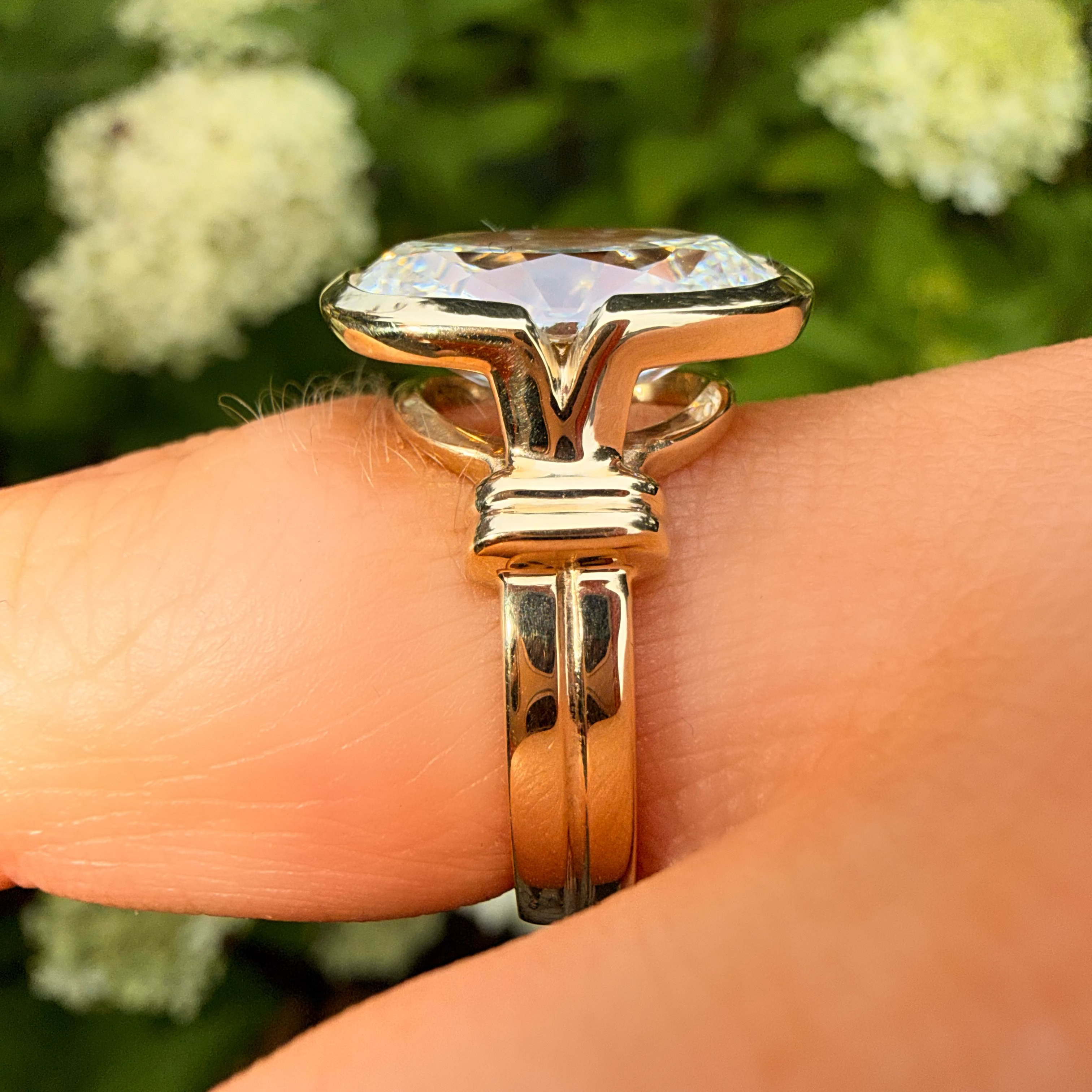 A gold ring with a large clear marquise-cut gemstone being worn on a finger, with a blurred green garden background and white flowers.
