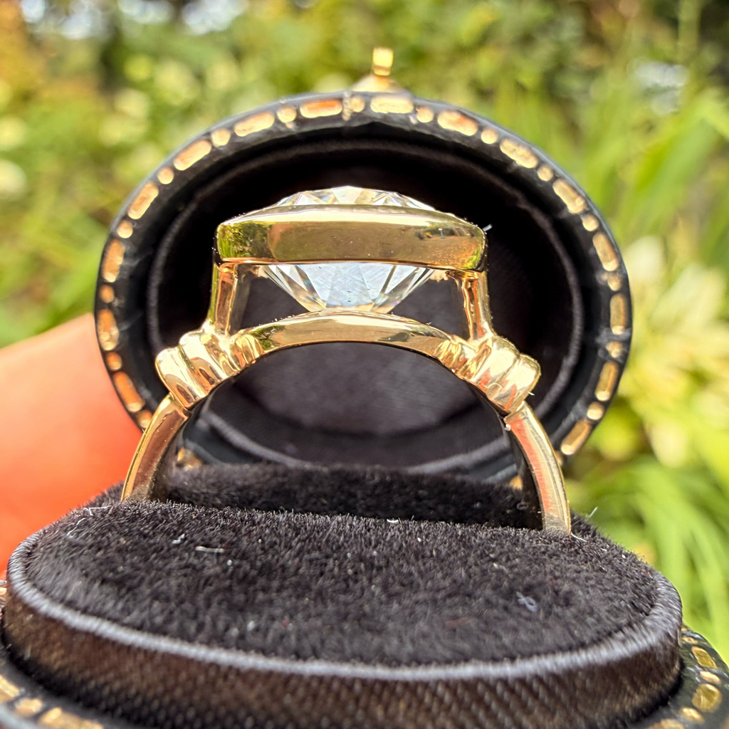 A gold ring with a large clear gemstone displayed inside an open black velvet ring box, with a blurred green outdoor background.