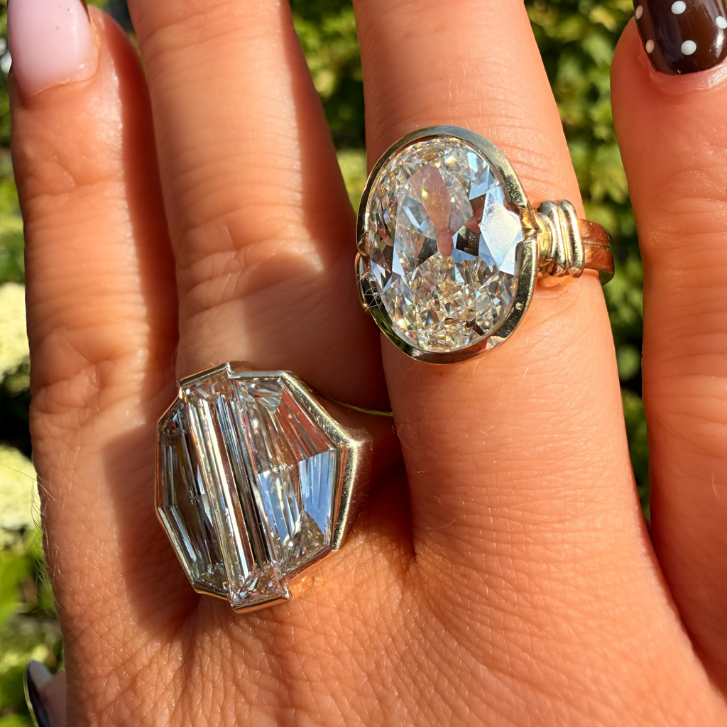 Two crystal rings on a hand with dark brown and white polka-dot nail polish, set against a blurred green outdoor background. One ring is a large oval clear crystal with a gold band, and the other is a faceted clear crystal with a silver-toned band.
