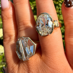 Two crystal rings on a hand with dark brown and white polka-dot nail polish, set against a blurred green outdoor background. One ring is a large oval clear crystal with a gold band, and the other is a faceted clear crystal with a silver-toned band.