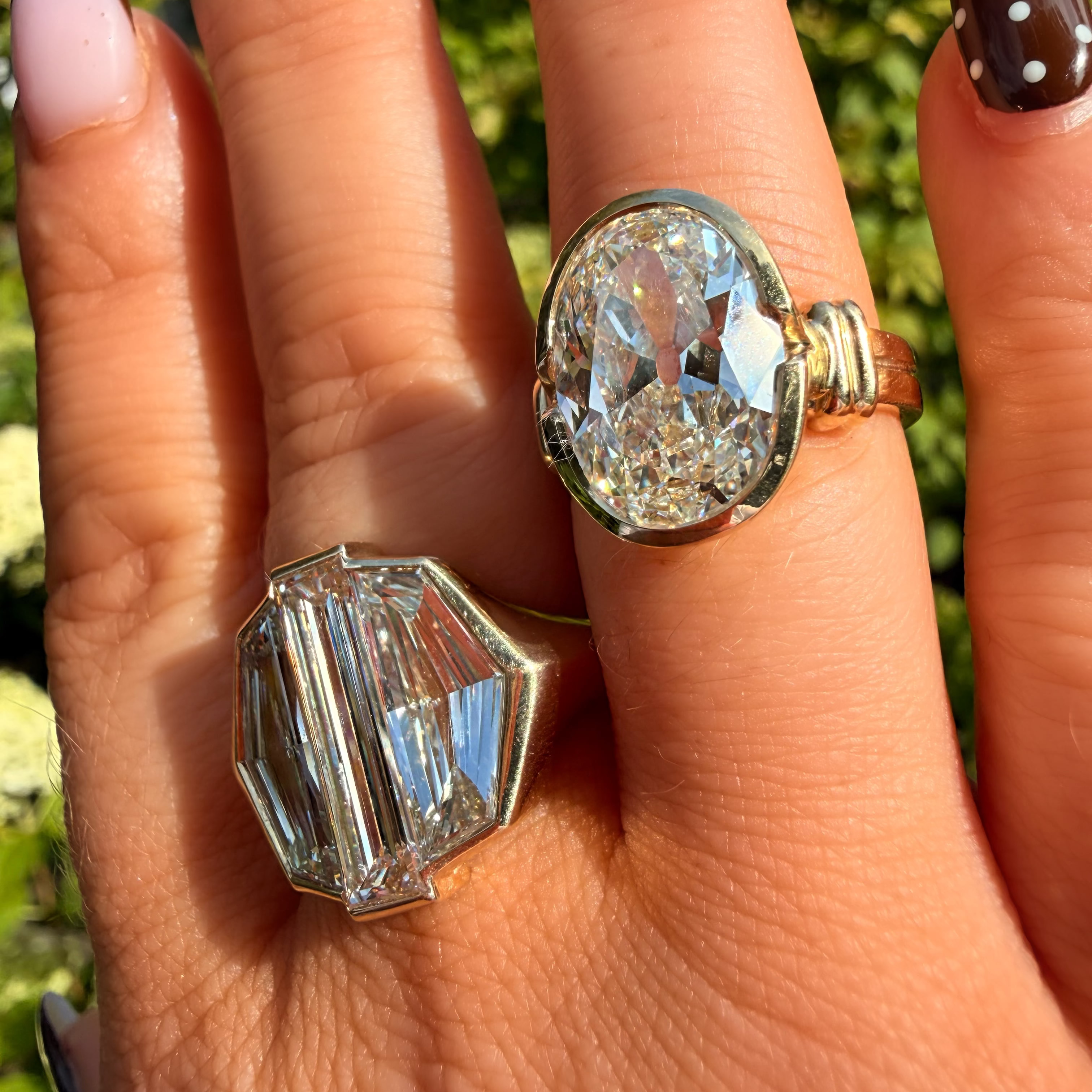 Two crystal rings on a hand with dark brown and white polka-dot nail polish, set against a blurred green outdoor background. One ring is a large oval clear crystal with a gold band, and the other is a faceted clear crystal with a silver-toned band.