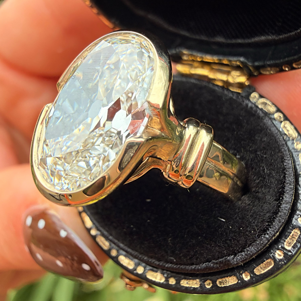 A close-up of a gold ring with a large oval clear gemstone displayed inside an open black ring box, held by a hand with brown nail polish against a blurred green outdoor background.