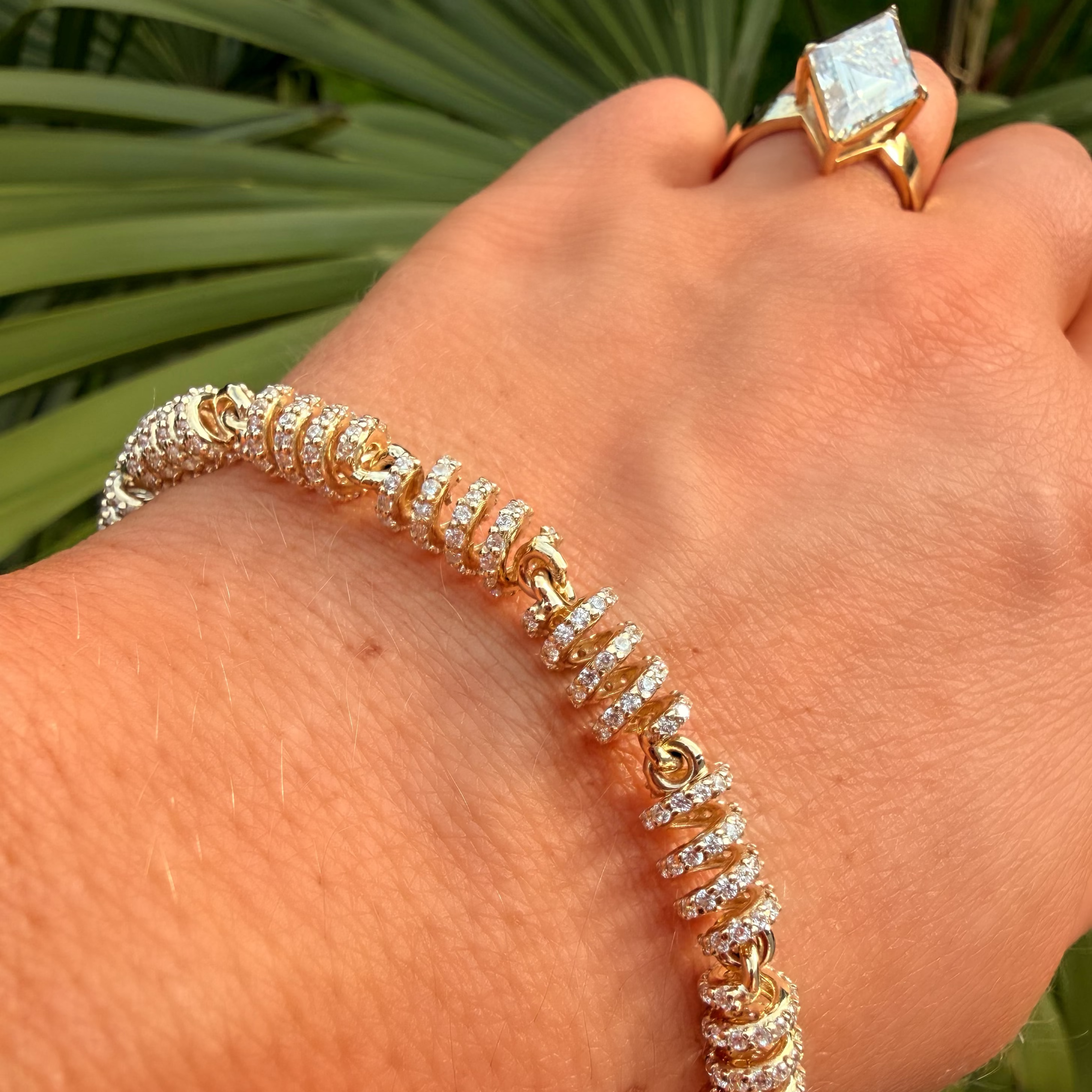 Hand wearing a gold and diamond bracelet with a matching ring against a green leafy background