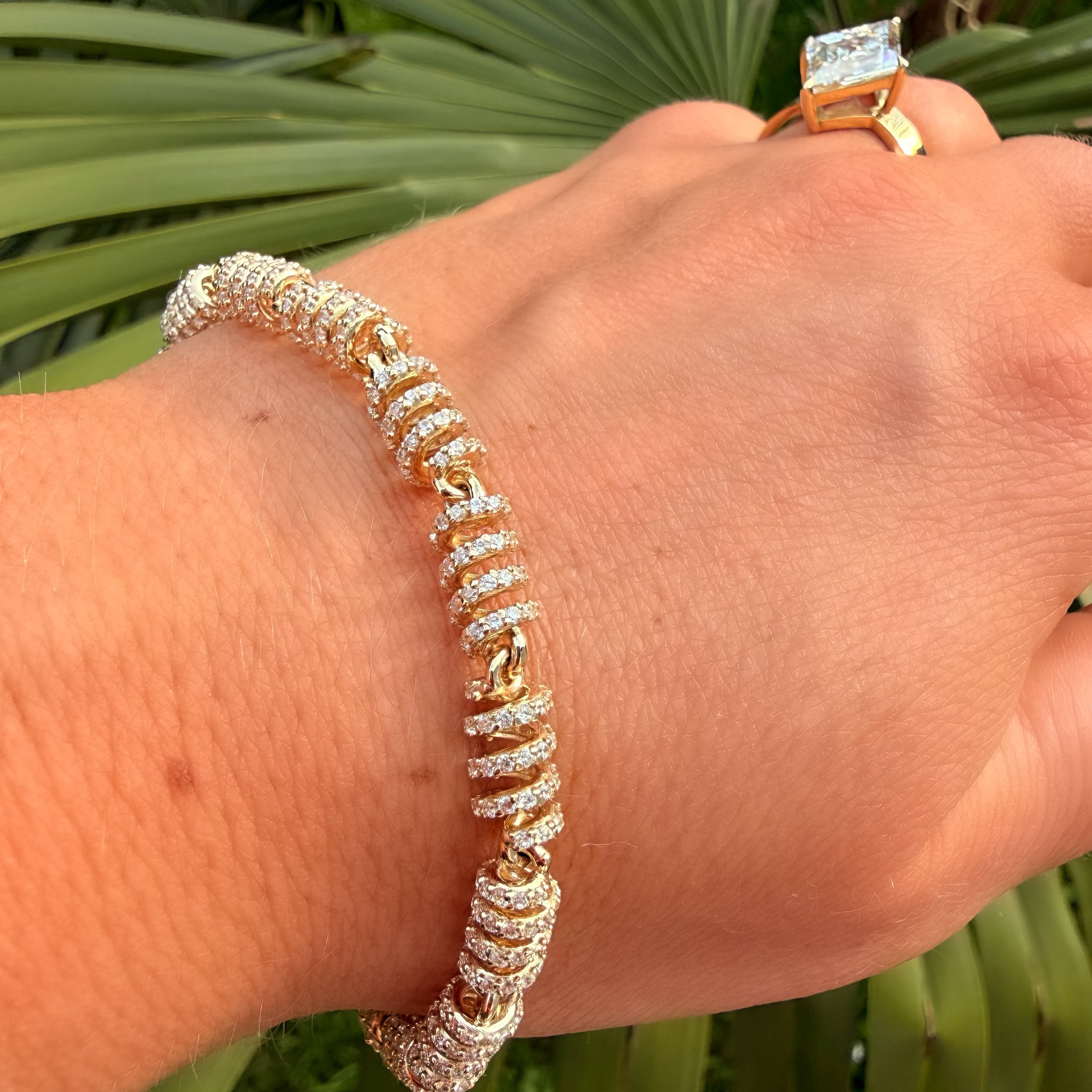 Hand wearing a diamond bracelet with a ring, against a green leafy background