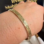 A textured gold bangle bracelet worn on a person's wrist, with a black jewelry box in the background showing gold lettering.