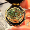 A close-up of a large, iridescent green marquise-cut gemstone set in a gold-toned octagonal ring box, held by a hand.