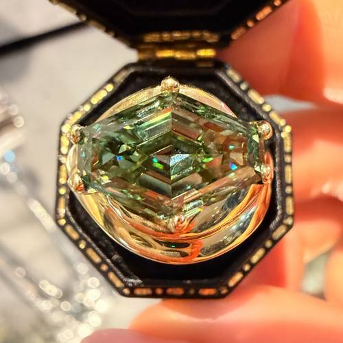 A close-up of a large, iridescent green marquise-cut gemstone set in a gold-toned octagonal ring box, held by a hand.