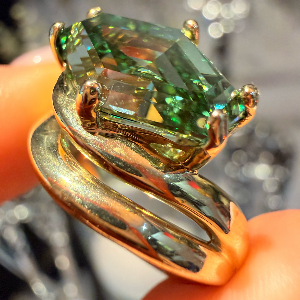 A close-up of a gold ring with a large iridescent green marquise-cut gemstone, held between fingers against a blurred reflective background.