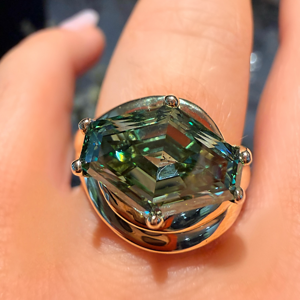 A close-up of a hand wearing a large ring with a faceted green gemstone in a gold-toned setting.