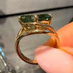 A close-up of a gold ring with a rectangular green gemstone, held by a hand with pink manicured nails against a blurred gray background.