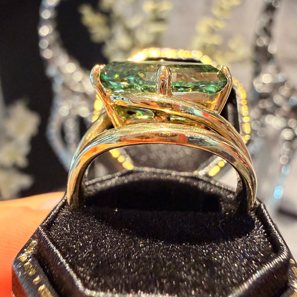 A gold ring with a rectangular green gemstone displayed in a black ring box, with a blurred jewelry setting in the background.