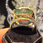A gold ring with a rectangular green gemstone displayed in a black ring box, with a blurred jewelry setting in the background.