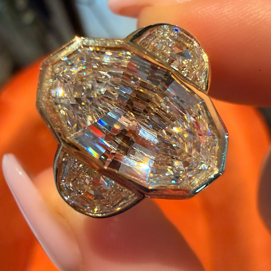 A close-up of a hand holding a large, ornate ring with a prominent emerald-cut center stone and smaller surrounding stones in a gold-toned setting, against an orange background.
