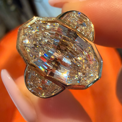 A close-up of a hand holding a large, ornate ring with a prominent emerald-cut center stone and smaller surrounding stones in a gold-toned setting, against an orange background.