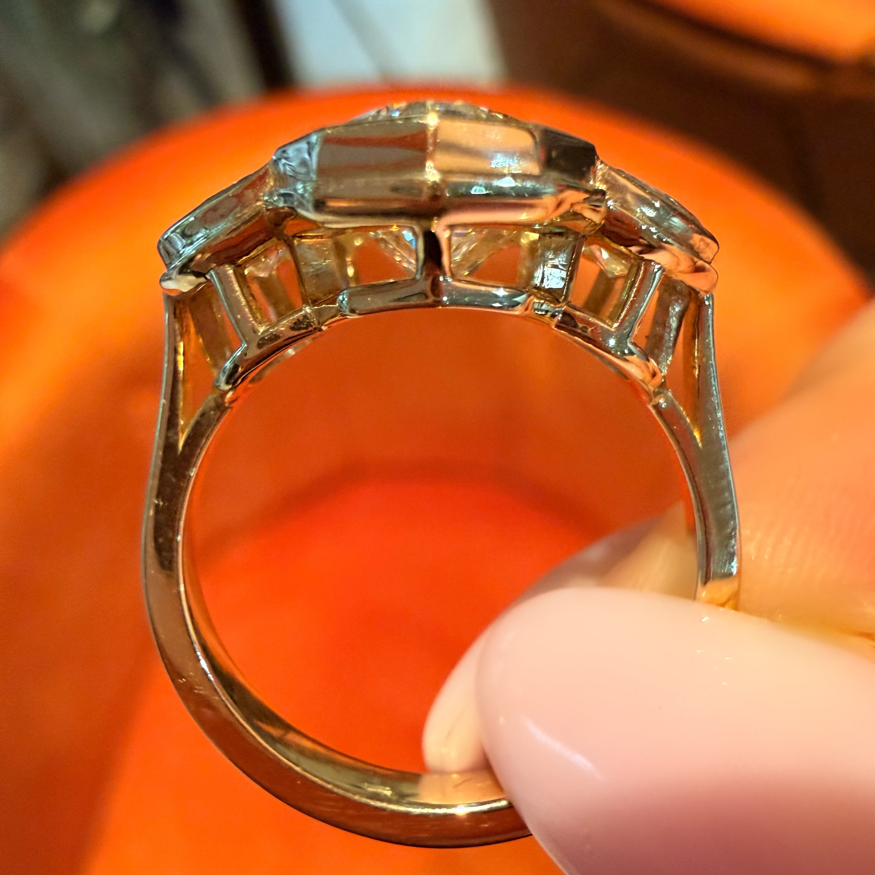 A close-up of a gold ring with a large rectangular clear gemstone held by a person's fingers against an orange background.