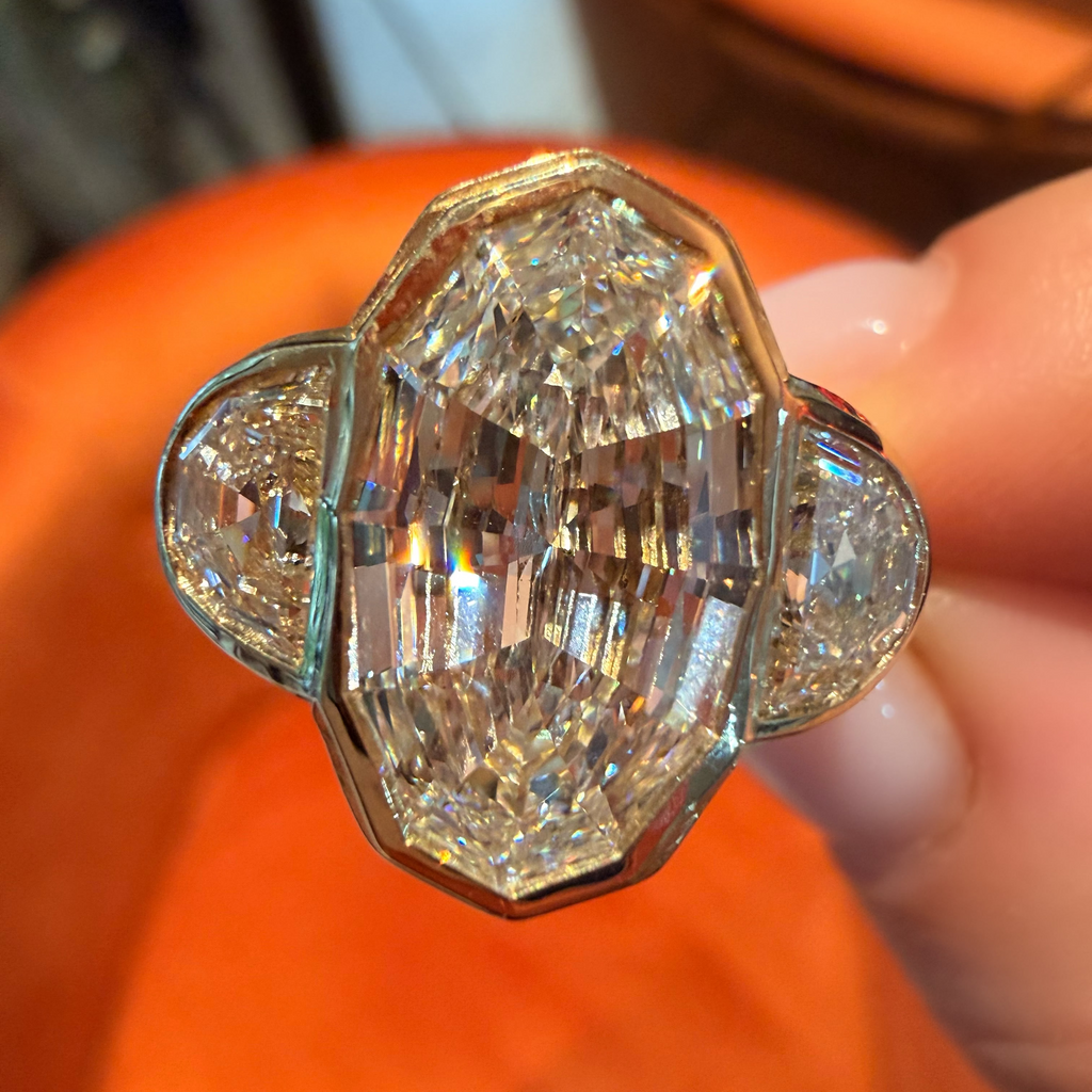 A close-up of a diamond ring held by fingers against a bright orange background. The ring features a large central marquise-cut diamond flanked by two smaller round diamonds in a gold setting.