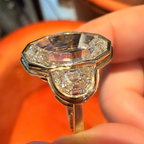 A close-up of a hand holding a vintage-style gold ring featuring a large, faceted clear gemstone and a smaller diamond accent, set against a bright orange background.