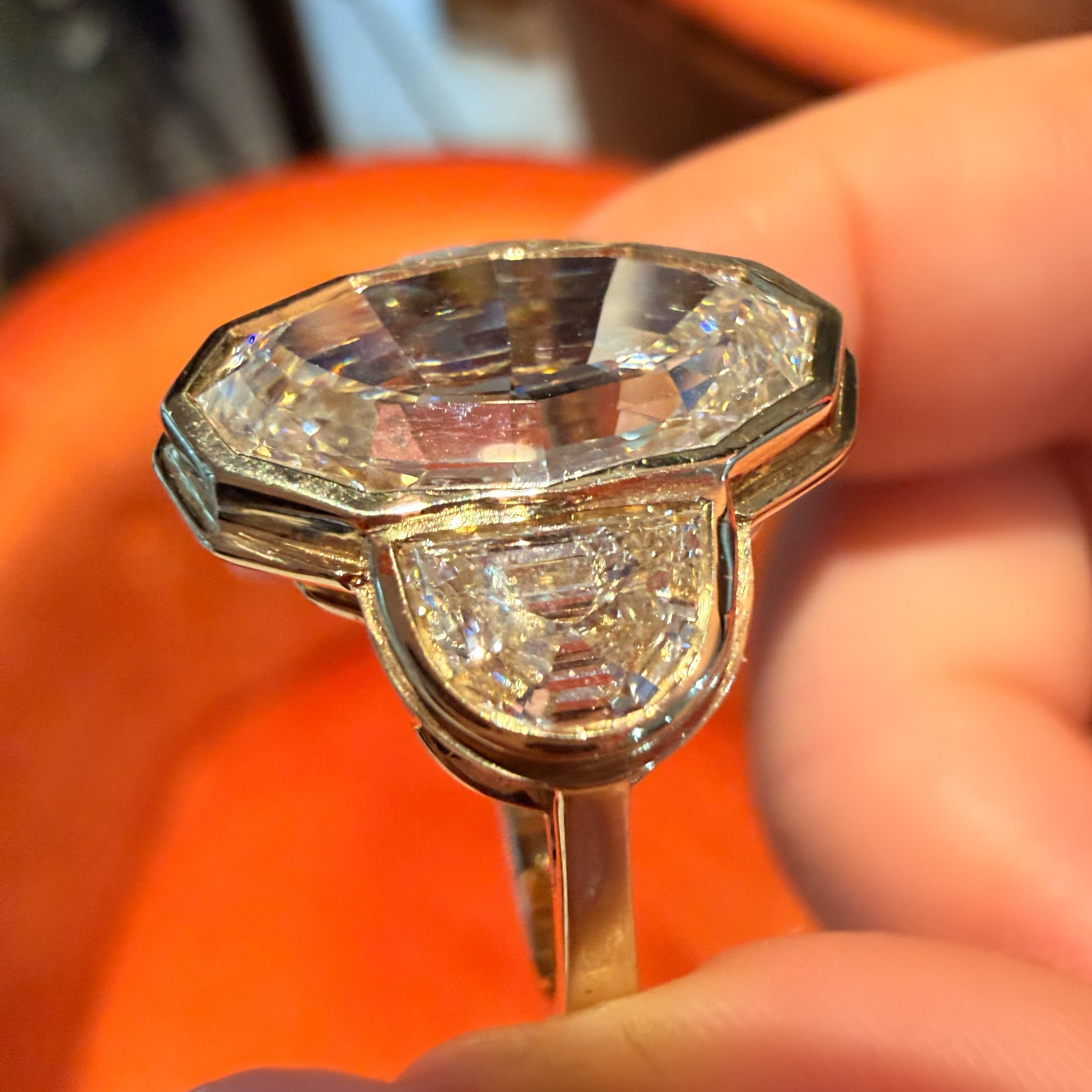 A close-up of a hand holding a vintage-style gold ring featuring a large, faceted clear gemstone and a smaller diamond accent, set against a bright orange background.