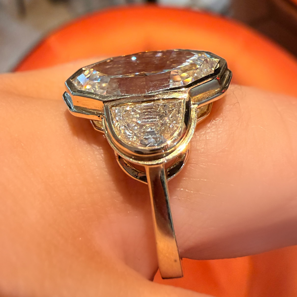 A close-up of a gold ring with a large clear faceted gemstone and smaller clear stones, worn on a finger against a blurred orange and indoor background.